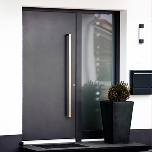 Image de catégorie pour portes d’entrée aluminium. On voit une porte d’entrée anthracite avec une poignée carrée argentée de l'extérieur avec un panneau latéral du côté de la serrure et un pot de fleurs à côté de l'entrée.
